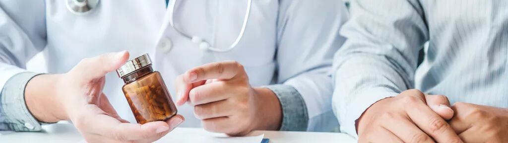 A medical professional holding and pointing to a bottle of medication for another person. There is a clipboard, pen, and medication blister pack in front of the medical professional