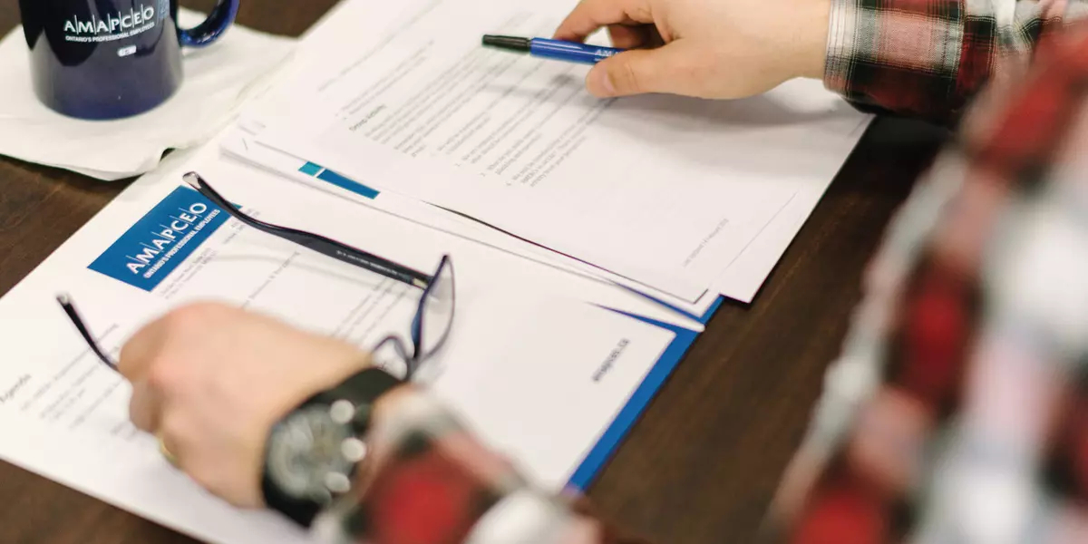 Hands of a member are placed over a number of AMAPCEO-branded papers. They are resting their glasses down with one hand and holding a pen with another.