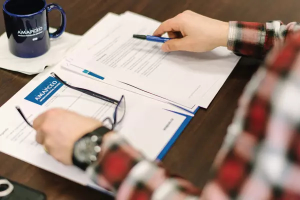 Hands of a member are placed over a number of AMAPCEO-branded papers. They are resting their glasses down with one hand and holding a pen with another.