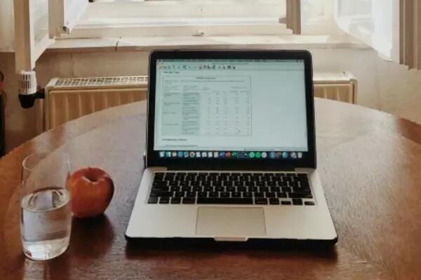 A laptop on a kitchen table with a glass of water and an apple next to it