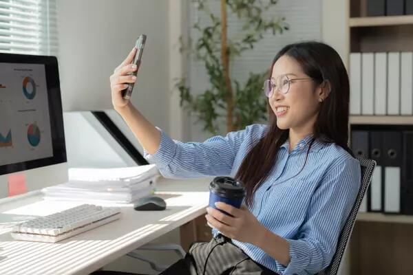 A person in a home office in front of a computer. They are recording a selfie with their phone in one hand. Their other hand is holding a coffee cup.