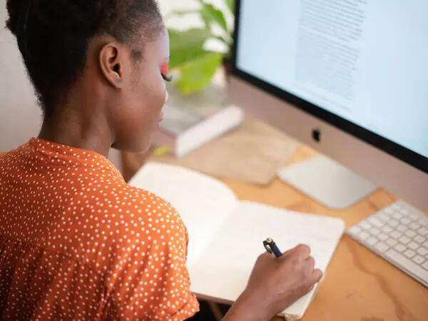 A person working from home, writing in a notebook and in front of a computer.