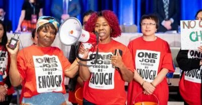 Picture of Toronto & York Region Labour Council President Andria Babbington speaking at the 2019 Annual Delegates' Conference accompanies by two colleagues wearing t-shirts that say One Job Should Be Enough.