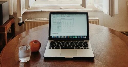 A laptop on a kitchen table with a glass of water and an apple next to it