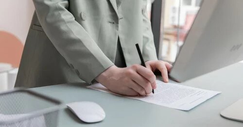 A person's hands taking notes on a paper with a monitor and mouse on the desk