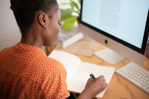 A person working from home, writing in a notebook and in front of a computer.