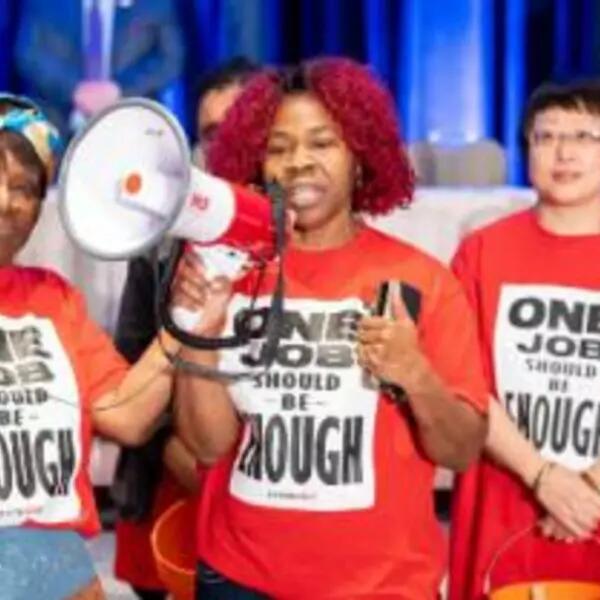 Picture of Toronto & York Region Labour Council President Andria Babbington speaking at the 2019 Annual Delegates' Conference accompanies by two colleagues wearing t-shirts that say One Job Should Be Enough.